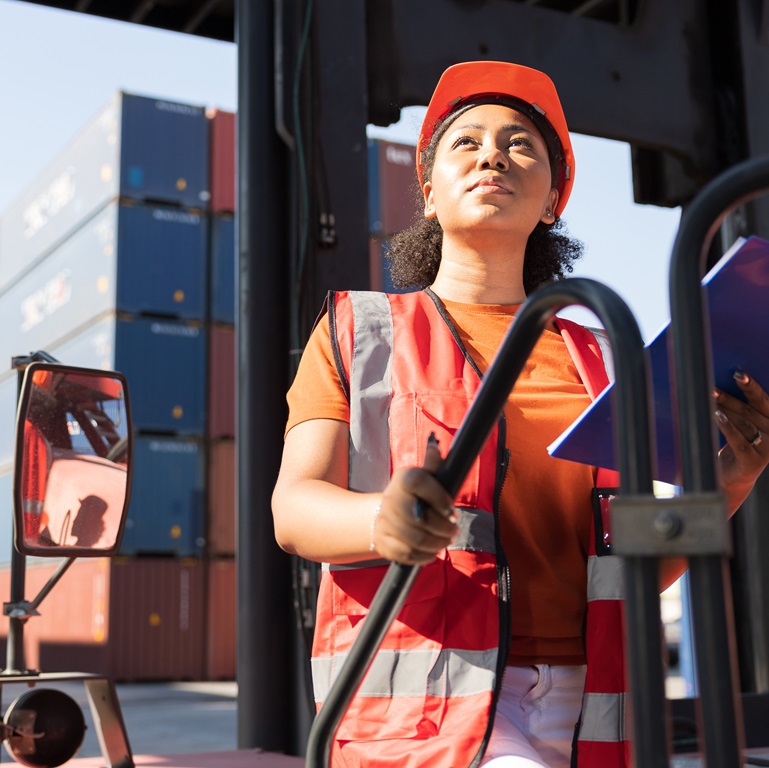 young female African factory worker or engineer holding clipboard and looking up to something on electric forklift trucks in 